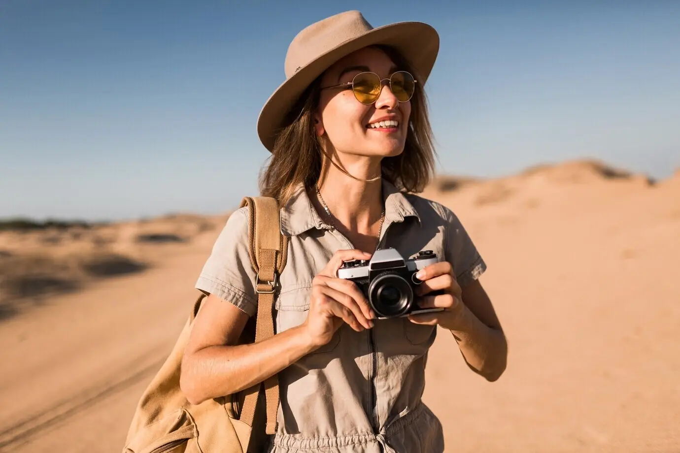 An attractive, stylish young woman in a khaki dress in a desert, traveling in Africa on safari, wearing a hat and a backpack, taking a photo with a vintage camera.