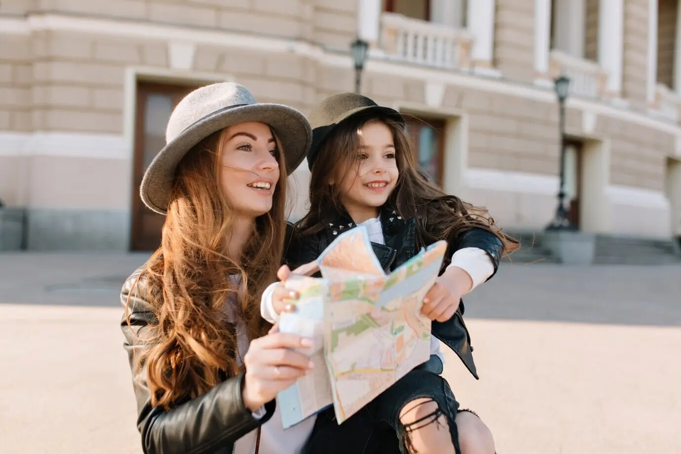A wonderful, inspired, curly-haired woman in a hat holds her pretty daughter and a city map, looking away. Outdoor portrait of two girls traveling around a new place and searching for beautiful sights.